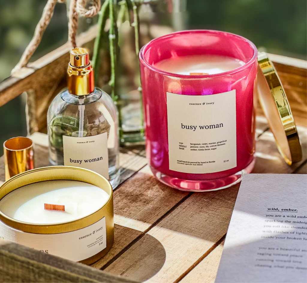 Two candles labeled 'busy woman' on a wooden surface with a bottle of perfume.