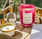Two candles labeled 'busy woman' on a wooden surface with a bottle of perfume.